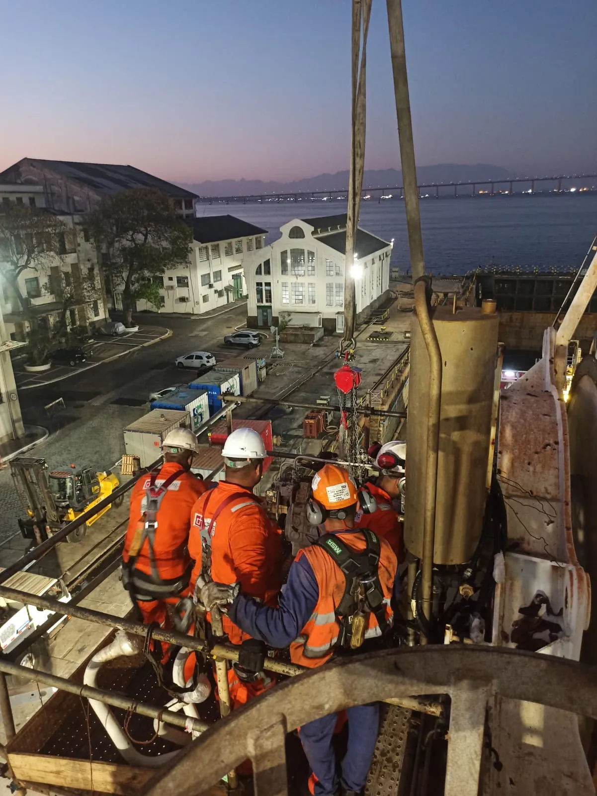 Equipe de técnicos em uma plataforma elevada ao amanhecer, com o porto e uma ponte ao fundo