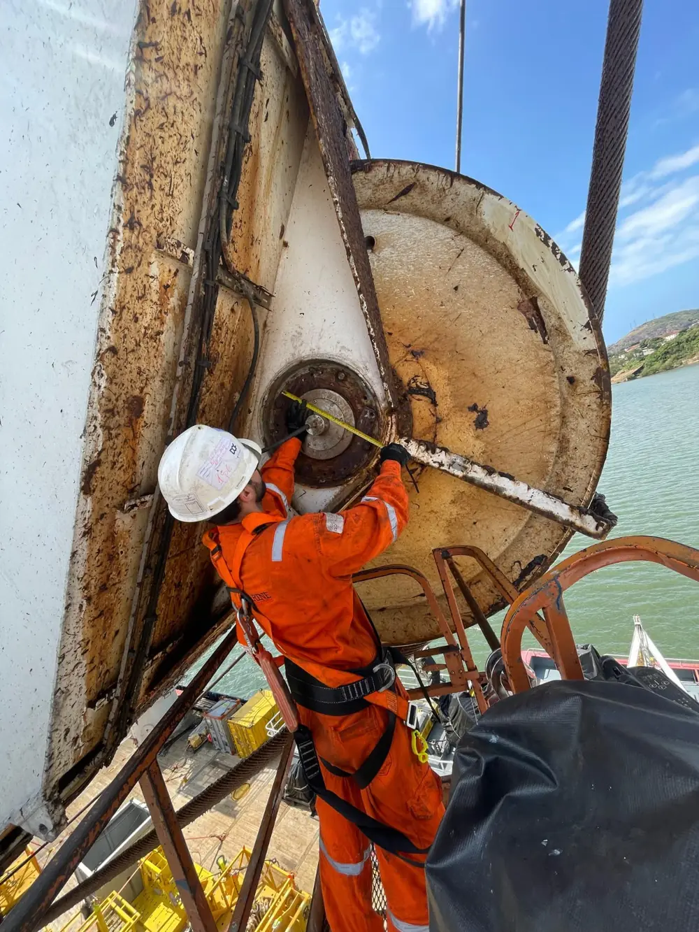 Técnico preso por cinto de segurança mede o cubo de um grande tambor enferrujado, com o mar ao fundo