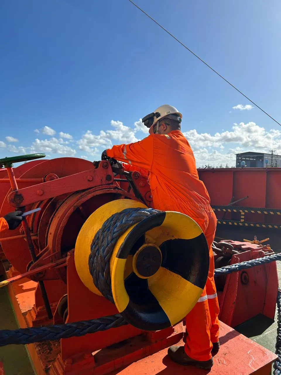 Técnico de macacão laranja trabalha em um guincho de amarração com cabo azul, no convés, sob céu azul